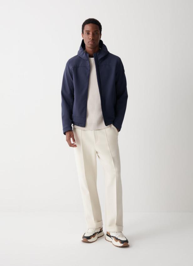 Ein dunkelhäutiger Mann in einer marineblauen Jacke, einem cremefarbenen Pullover und hellen Hosen steht vor einem weißen Hintergrund.