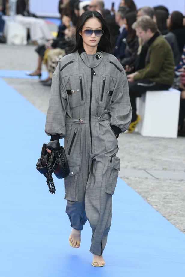 Eine barfüßige Frau in einem grauen Overall mit Sonnenbrille und Handtasche auf einem Laufsteg.