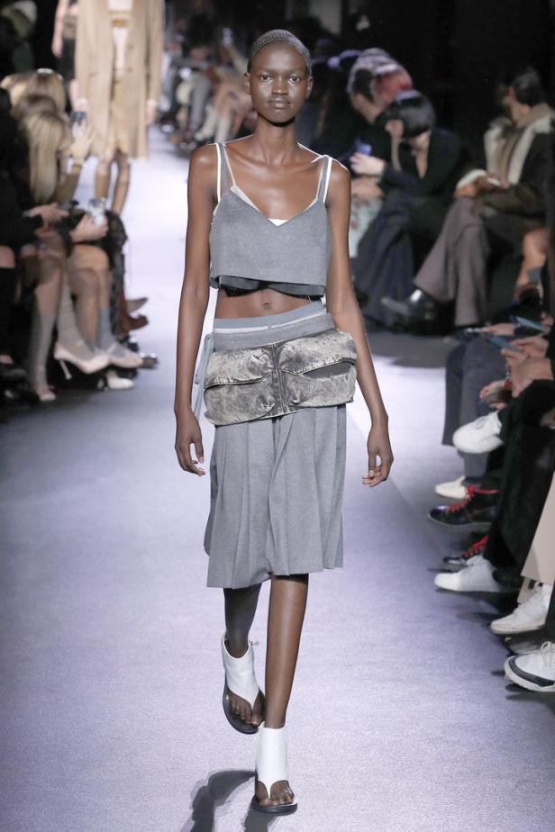 Eine dunkelhäutige Frau in einem grauen zweiteiligen Outfit und weißen Sandalen auf einem Laufsteg.