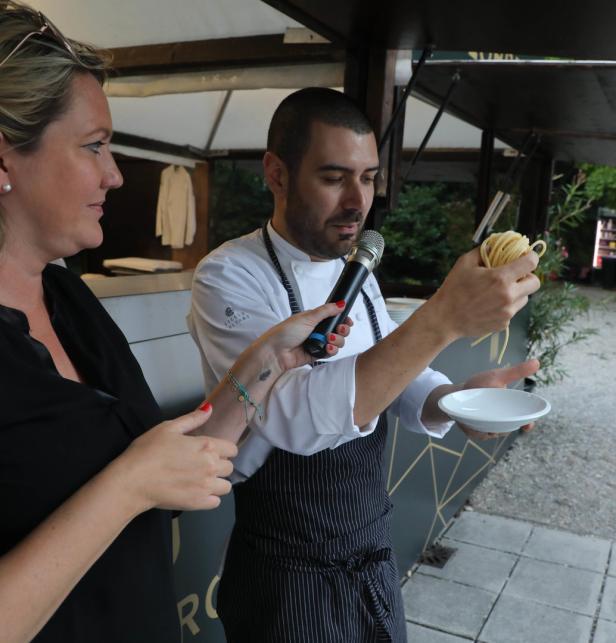 Ein Koch präsentiert Nudeln in einem Restaurant, während eine Frau ihm ein Mikrofon hält.