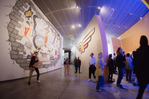 Besucher betrachten eine Ausstellung mit Wandmalereien und einem geflügelten Logo.
