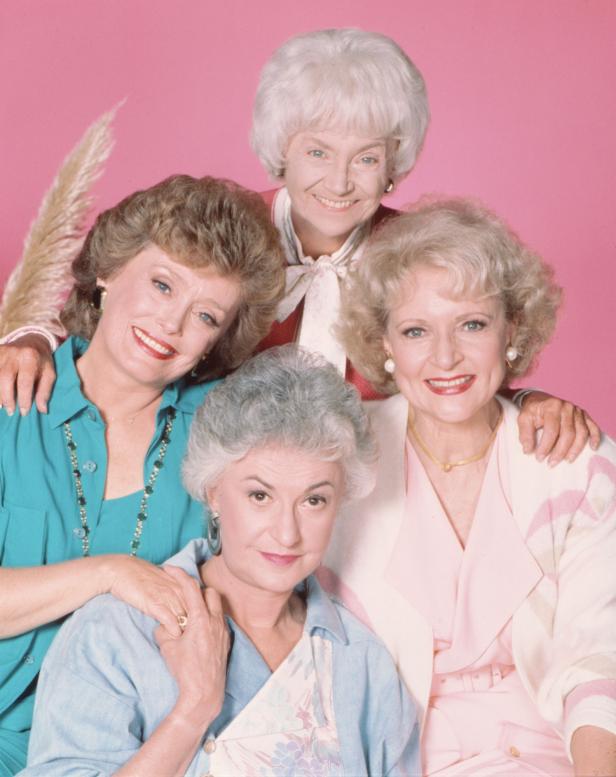 Die vier Hauptdarstellerinnen der Fernsehserie „Golden Girls“ posieren vor einem rosa Hintergrund.