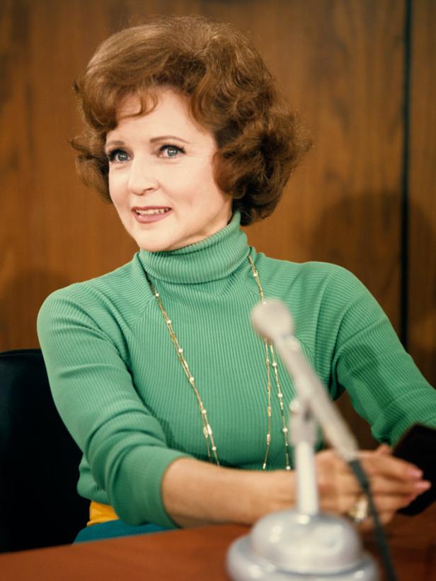 Betty White sitzt an einem Tisch mit einem Mikrofon und lächelt.