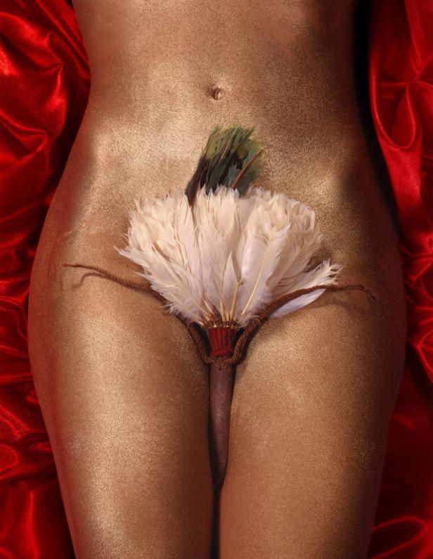 Der goldene Körper einer Frau, bedeckt mit einem Fächer aus Federn, liegt auf rotem Stoff.