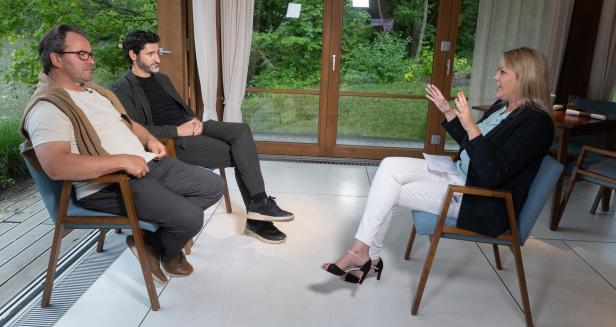 Eine Frau interviewt zwei Männer in einem Raum mit Blick auf die Natur.