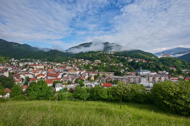 Blick auf eine Stadt in einem Tal, umgeben von bewaldeten Hügeln und Wolken.