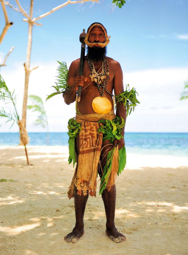 Ein Mann in traditioneller Kleidung steht am Strand mit einer Keule in der Hand.