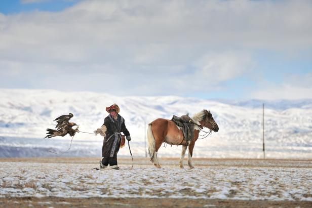 Ein kasachischer Adlerjäger mit seinem Steinadler und Pferd in der verschneiten Steppe.