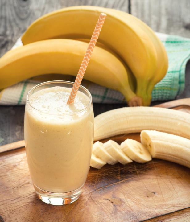Ein Glas Bananen-Smoothie mit Strohhalm steht neben Bananen auf einem Holzbrett.