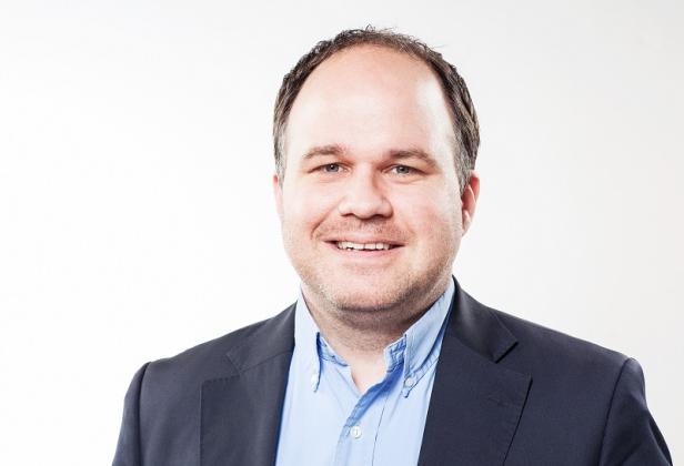Ein lächelnder Mann in einem blauen Hemd und einem dunklen Sakko vor einem weißen Hintergrund.