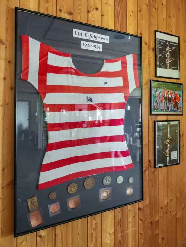 Ein gerahmtes Trikot mit roten und weißen Streifen und Medaillen darunter an einer Holzwand.