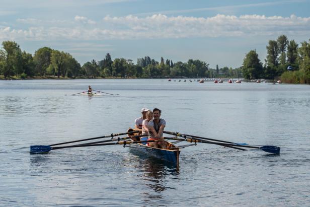 Drei Personen rudern in einem blauen Ruderboot auf einem See.
