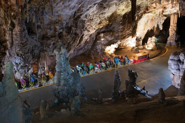 Eine Touristengruppe fährt mit einer kleinen Bahn durch eine beleuchtete Höhle mit Stalaktiten und Stalagmiten.