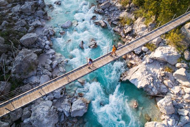Zwei Personen überqueren eine Hängebrücke über einen türkisfarbenen Fluss.