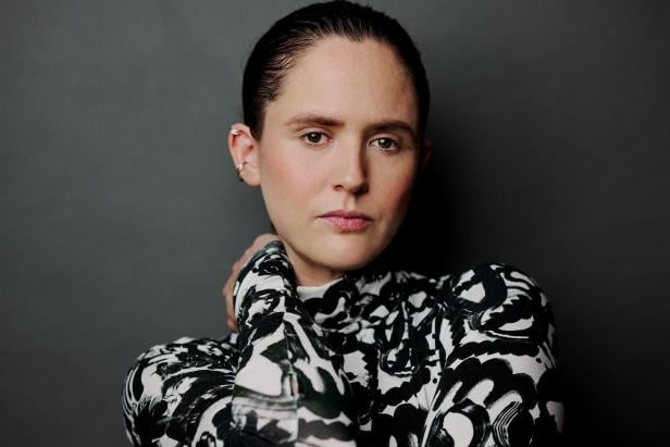 Eine Frau mit zurückgekämmten Haaren und einem schwarz-weiß gemusterten Oberteil posiert vor einem grauen Hintergrund.