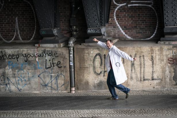 Ein Mann im weißen Arztkittel tanzt vor einer Wand mit Graffiti.