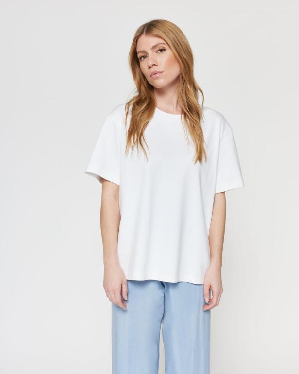 Eine Frau mit blonden Haaren trägt ein weißes T-Shirt und eine hellblaue Hose.