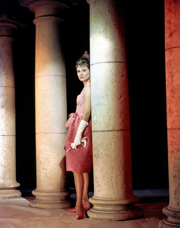 Audrey Hepburn in einem rosa Kleid, posierend zwischen Säulen.