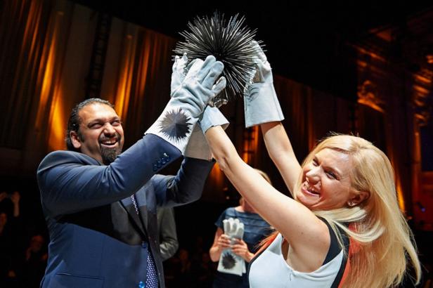 Zwei Personen mit Handschuhen halten eine stachelige Kugel hoch und lächeln.