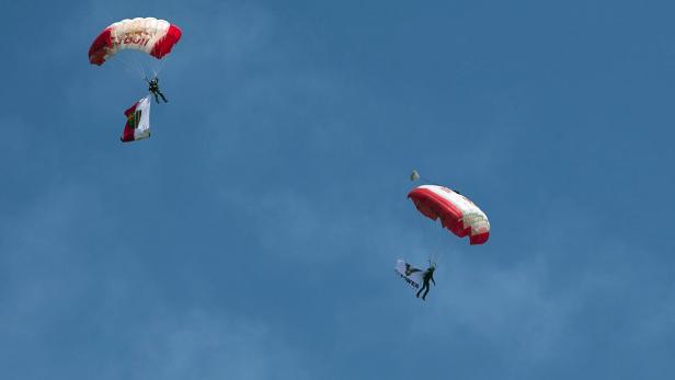 Zwei Fallschirmspringer mit rot-weißen Fallschirmen und Flaggen am Himmel.