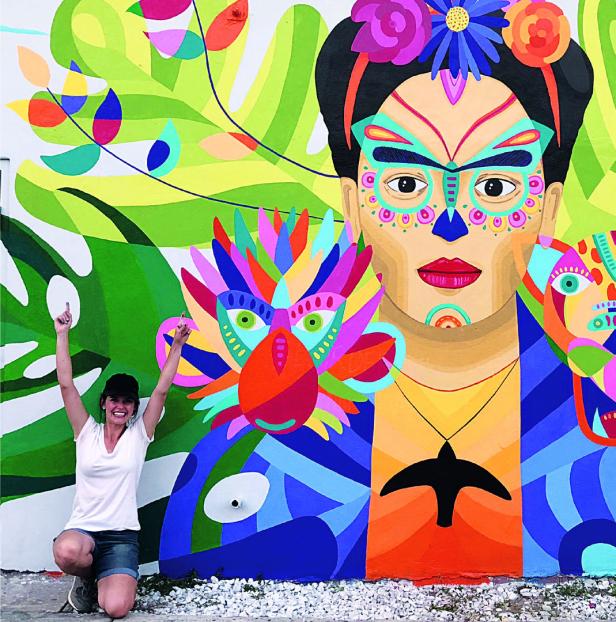 Eine Frau posiert vor einem farbenfrohen Wandgemälde, das Frida Kahlo darstellt.