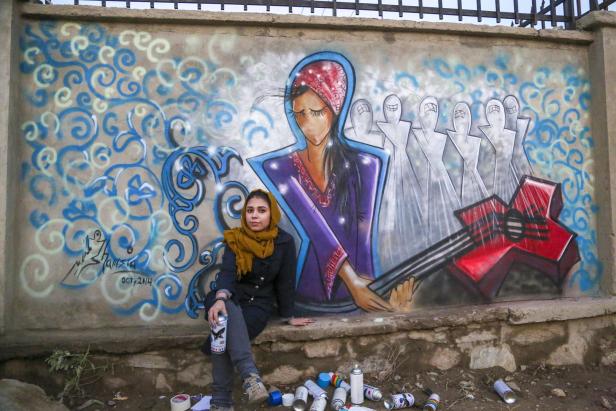 Eine junge Frau sitzt vor einem Graffiti mit einer stilisierten Frauengestalt und Geisterfiguren.