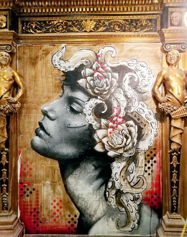 Ein Porträt einer Frau mit Tentakeln und Blumen im Haar, umrahmt von goldenen Verzierungen.