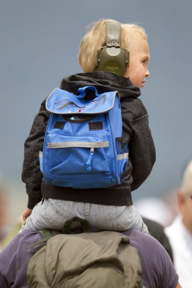 Ein Junge mit Kopfhörern sitzt auf den Schultern eines Erwachsenen und trägt einen blauen Rucksack.