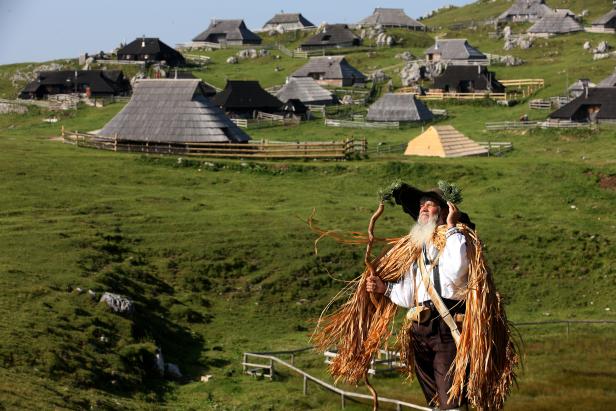 Ein Mann in Tracht steht vor einer Almhütte-Siedlung.