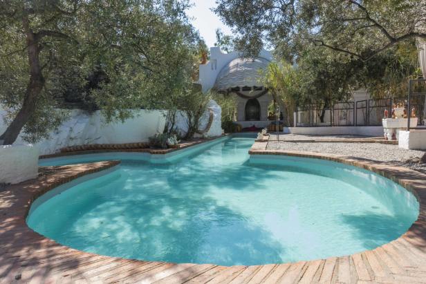Ein türkisfarbener Pool mit Backsteineinfassung vor einem weißen Gebäude und Olivenbäumen.