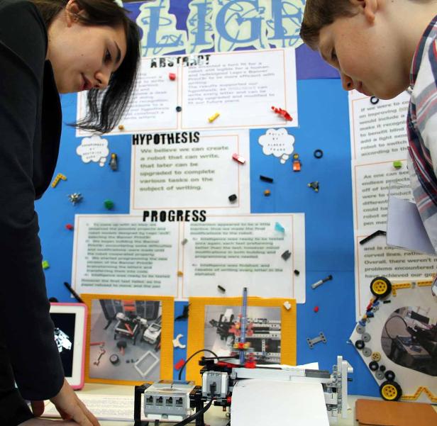 Zwei Jugendliche betrachten einen Projektstand mit einem schreibenden Lego-Roboter.