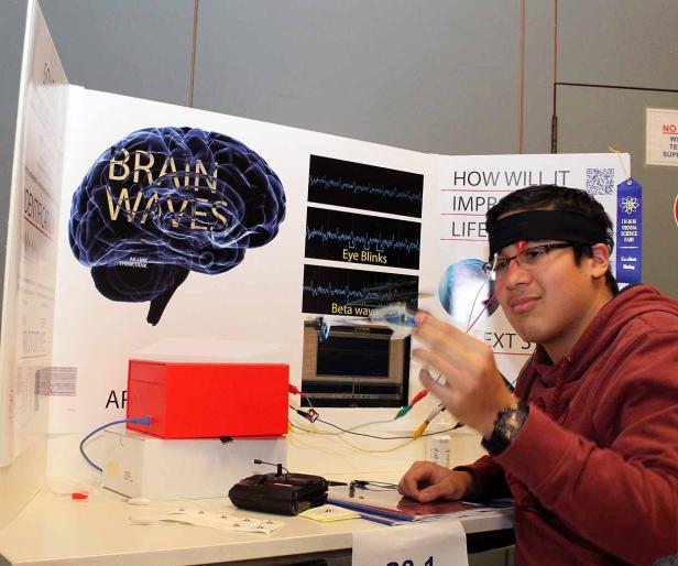 Ein junger Mann mit Elektroden am Kopf präsentiert ein Projekt über Gehirnwellen auf einer Wissenschaftsmesse.