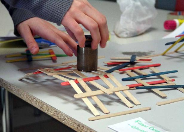 Eine Person testet die Stabilität einer Brücke aus Eisstielen mit einem Gewicht.