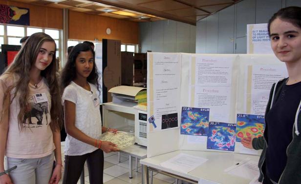 Drei junge Frauen stehen vor einem Tisch mit einem wissenschaftlichen Projekt über Mikrowellen.