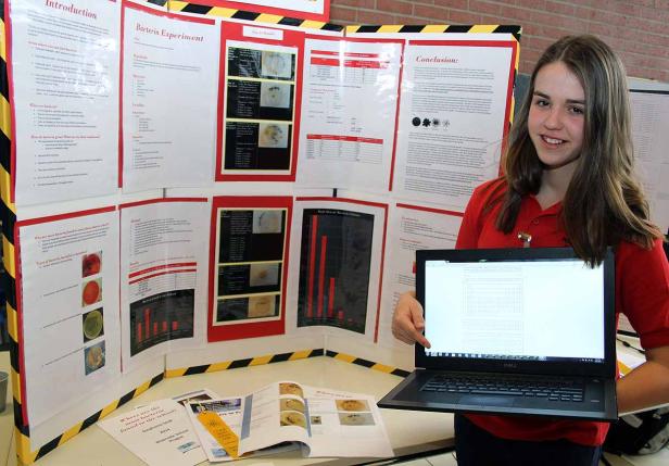 Ein Mädchen präsentiert ihr Bakterien-Experiment mit Laptop und Schaubild.