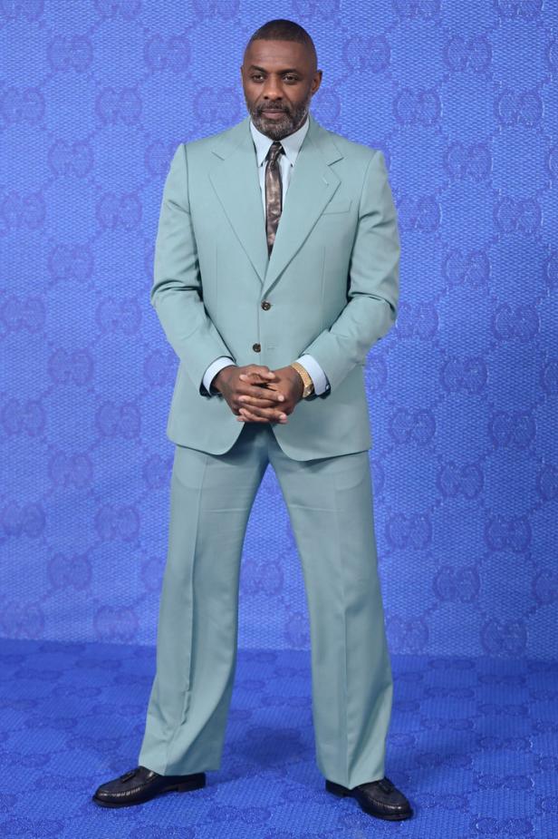 Idris Elba trägt einen hellgrünen Anzug vor einem blauen Hintergrund mit Gucci-Logos.