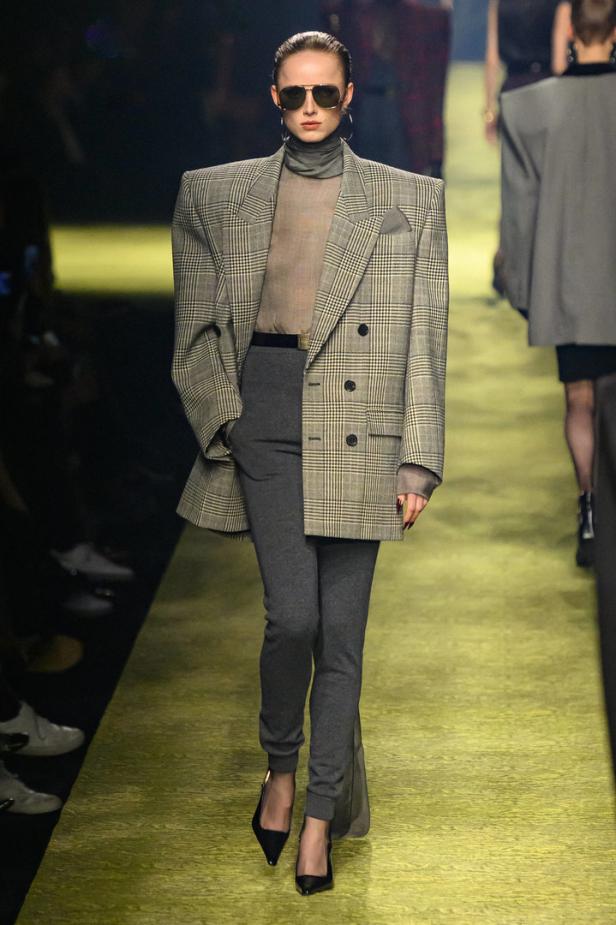 Eine Frau mit Sonnenbrille präsentiert ein Outfit mit übergroßem Blazer auf dem Laufsteg.