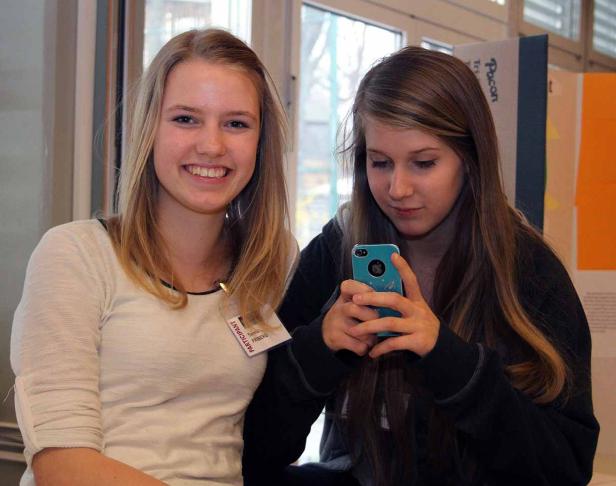 Zwei junge Frauen, eine lächelnd, die andere auf ihr Smartphone schauend.