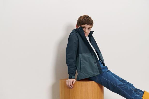 Ein Junge sitzt auf einem Holzhocker und trägt eine blaugrüne Jacke und Jeans.