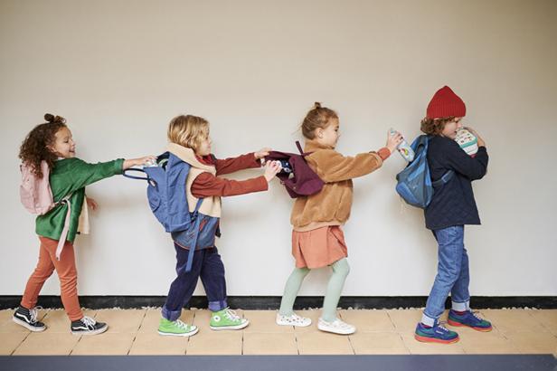Vier Kinder mit Rucksäcken gehen hintereinander und halten sich an den Rucksäcken fest.