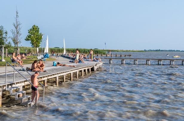 Das Ufer des Neusiedler Sees, Menschen liegen auf dem Holzsteg, ein Mann in Badehose geht ins Wasser