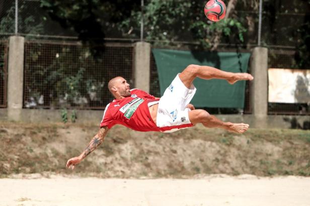 Fußballspieler im Sand in seitlicher Lage visiert den Ball an und tritt