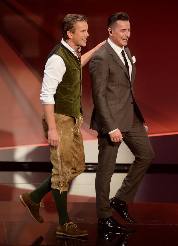 Zwei Männer, einer in Tracht, der andere im Anzug, lachen und gehen nebeneinander.