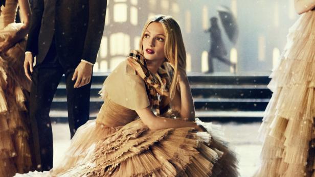 Eine blonde Frau sitzt in einem goldenen Kleid mit Schal vor einer verschneiten Kulisse.