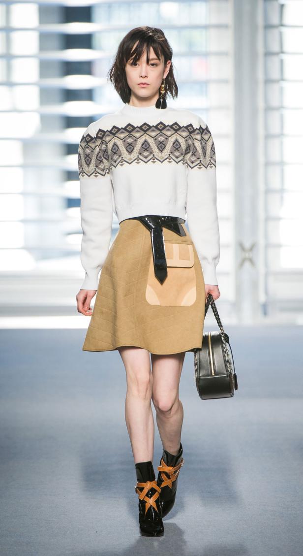 Eine Frau präsentiert einen weißen Pullover mit Muster, einen beigen Rock und schwarze Stiefel auf dem Laufsteg.