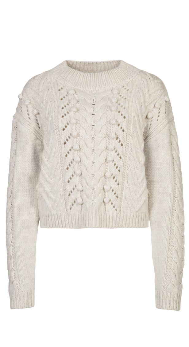 Ein cremefarbener, kurzer Strickpullover mit Zopfmuster und kleinen Bommeln.