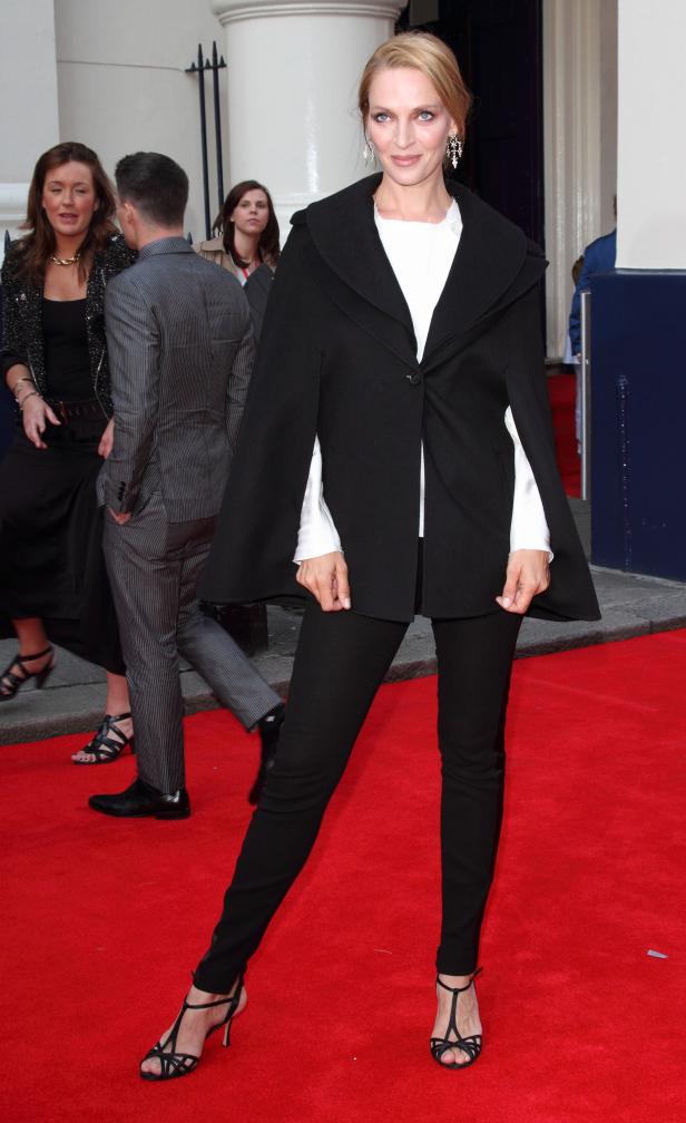 Uma Thurman posiert auf einem roten Teppich in einem schwarzen Cape und schwarzen Hosen.