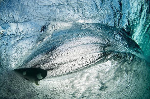 Ein Surfbrett ist unter einer Welle im türkisfarbenen Wasser zu sehen.