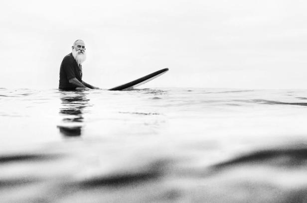 Ein älterer Mann mit Bart sitzt auf einem Surfbrett im Wasser.