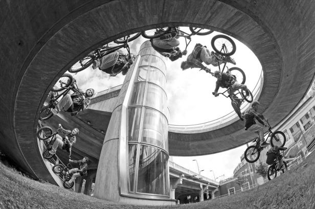 Mehrere BMX-Fahrer führen Tricks unter einer Betonbrücke und um einen gläsernen Aufzugsschacht herum aus.